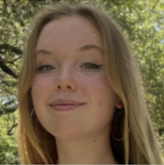 A young woman with long blonde hair and freckles smiling for a headshot. She is wearing large hoop earrings, and the background shows blurred green trees in natural sunlight.
