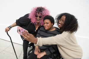 Three woman take a selfie together. The woman on the left has a cane in her hand. The woman is sitting in a wheelchair. The woman on the right is holding the phone away to take the selfie.