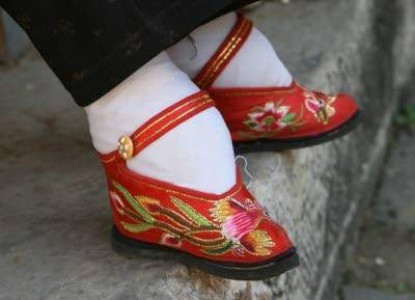 A woman’s bound feet from the ankle down, wearing white socks and very small red shoes with bright embroidered flowers, leaves and a bird.
