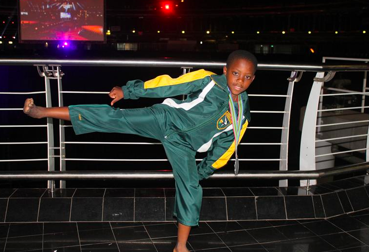 The same boy as above, now wearing a green martial arts uniform with a medal hanging from his neck. He is standing on his left leg while kicking his right leg out in a martial arts move. He is looking right at the camera with a stadium and large flat screen monitor behind him.