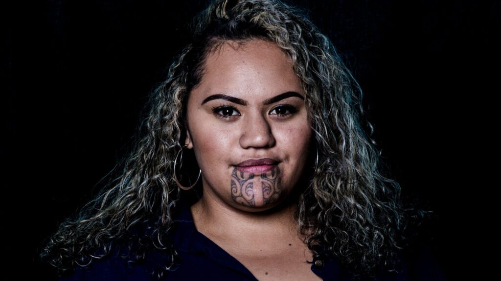 A Maori woman with olive-toned skin, dark eyebrows, long, wavy, curly, salt and pepper hair with blue highlights that falls past her shoulders. The woman has a Moko–a blue ink tattoo from the bottom of her bottom lip to just under her chin. The tattoo has traditional Maori carving patterns with thick lines and symmetrical swirls. She is looking right at the camera with a closed-mouth smile.
