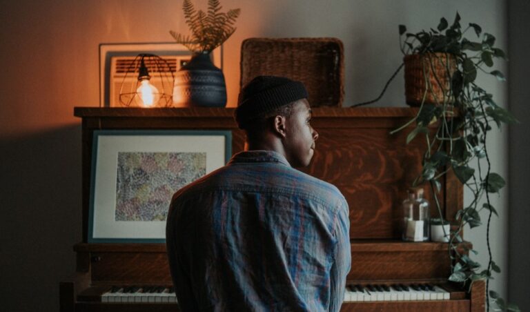 teenage person with a dark brown skin tone sitting in front of a piano and looking out the right-side window