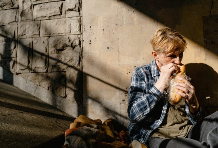 A disheveled, middle-aged person sits in an alley eating a sandwich