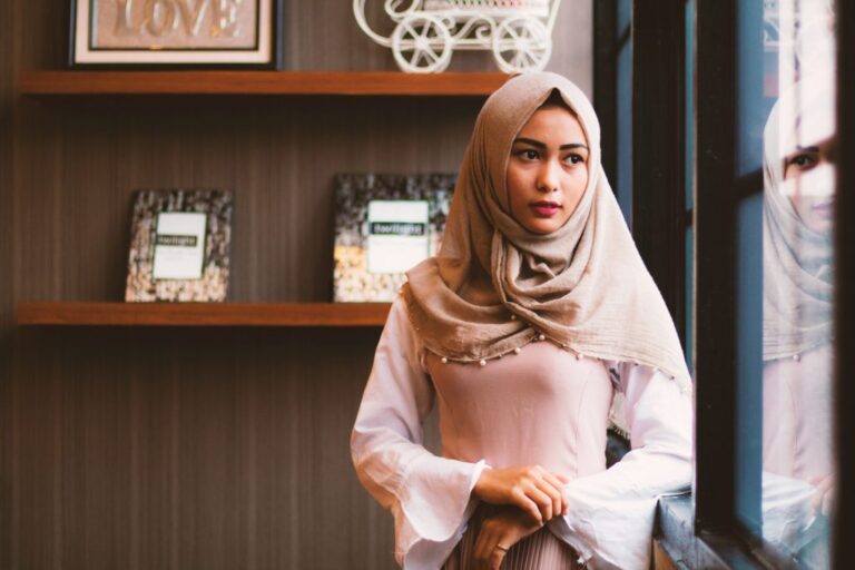 A South Asian young woman wearing a light pink dress with flowy sleeves, a cream colored hijab, looking out a window. Her reflection in the window is visible and behind her are brown wooden shelves with picture frames, and an art piece with the word 'Love'.]