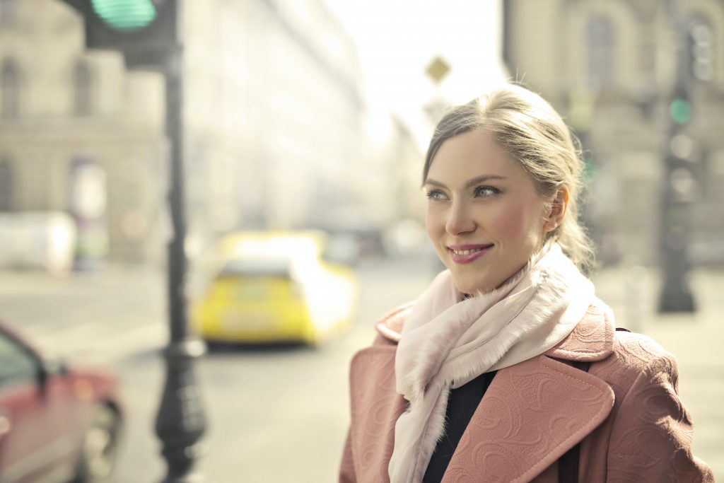 A white woman with straight blonde hair pulled back in a ponytail wearing a light pink scarf and a darker pink coat with a wide lapel. She is standing on a city sidewalk near a street with passing cars. She is smiling slightly and looking toward the street.