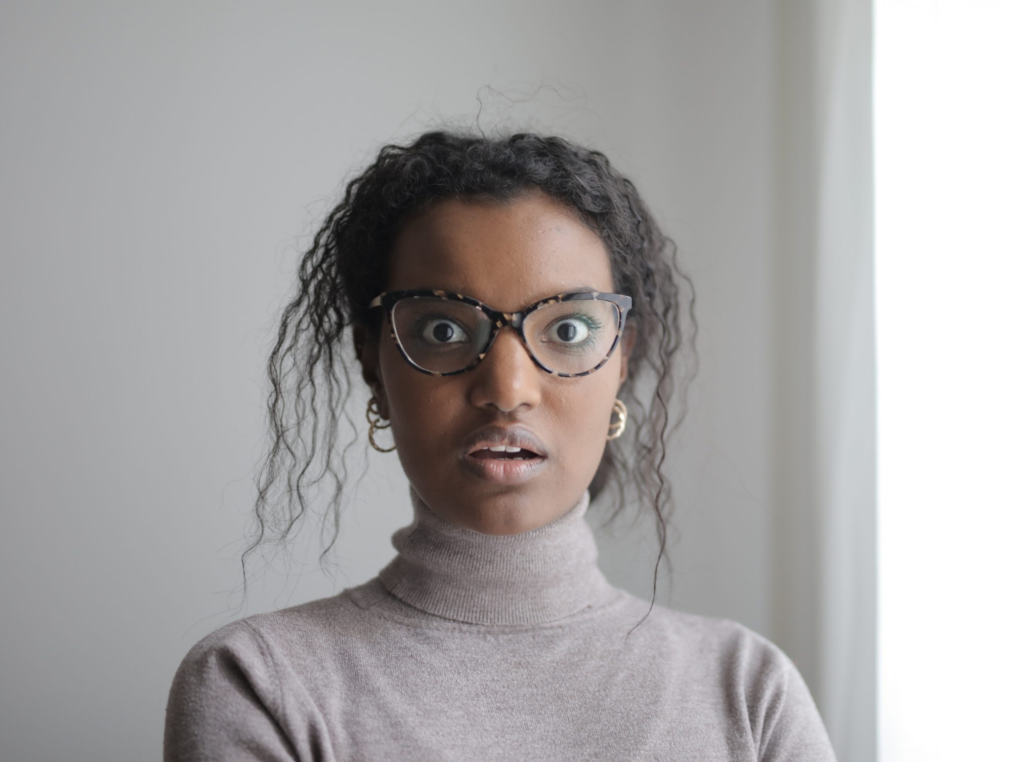 A young woman with dark skin, glasses, hoop earrings and a beige turtleneck looks directly at the camera with an expression of surprise and concern.]