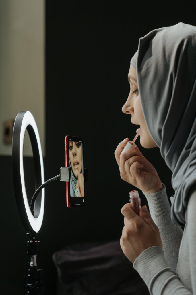 A woman wearing a gray hijab applying lip gloss in front of a round light. Her image can be seen also in her mobile phone that is connected to the light with a stand. She is using her phone camera either to record the makeup application or as a mirror.