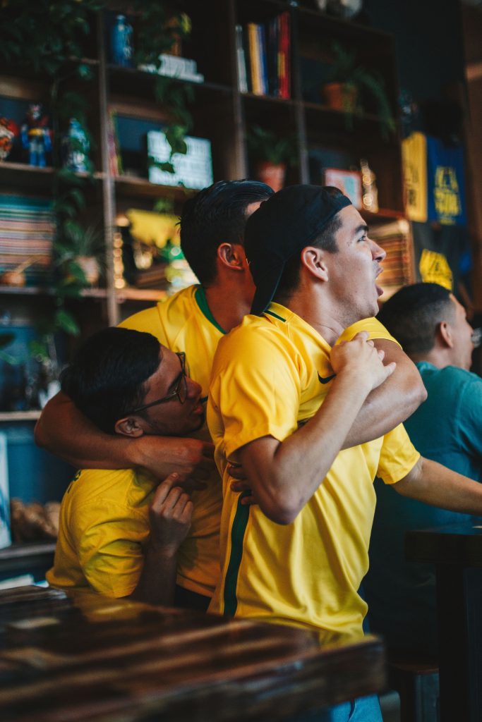 Three male soccer/football fans wearing matching yellow jerseys hug while watching the game in a bar/pub.