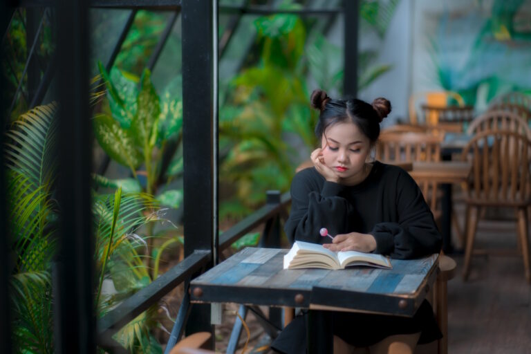 An Asian girl with two hair buns wearing a black sweater sits at a cafe reading a book on the table in front of her. Behind her, a window reveals green plants.