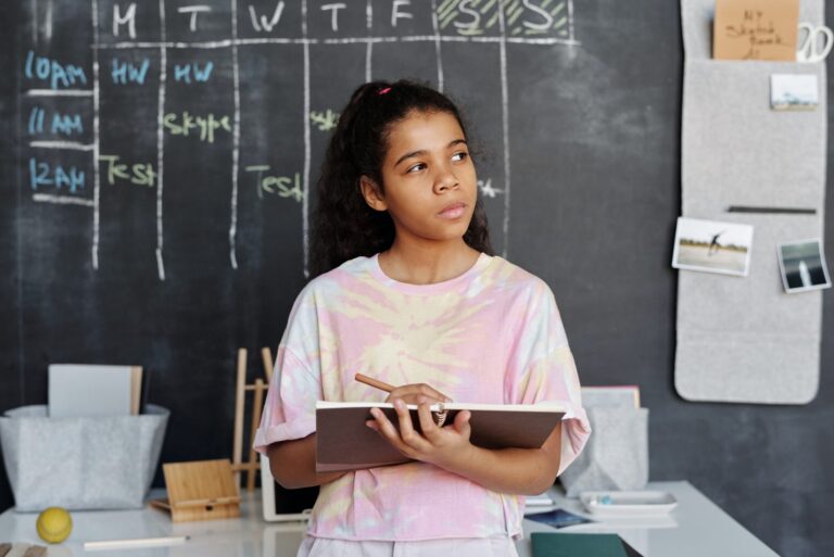 A teenage girl with light brown skin wearing a pink and white shirt. She is standing in front of a desk, holding a notepad and pen while looking upward to the left. Behind her is a chalkboard with a Monday to Friday schedule noting tests and Skype sessions.]