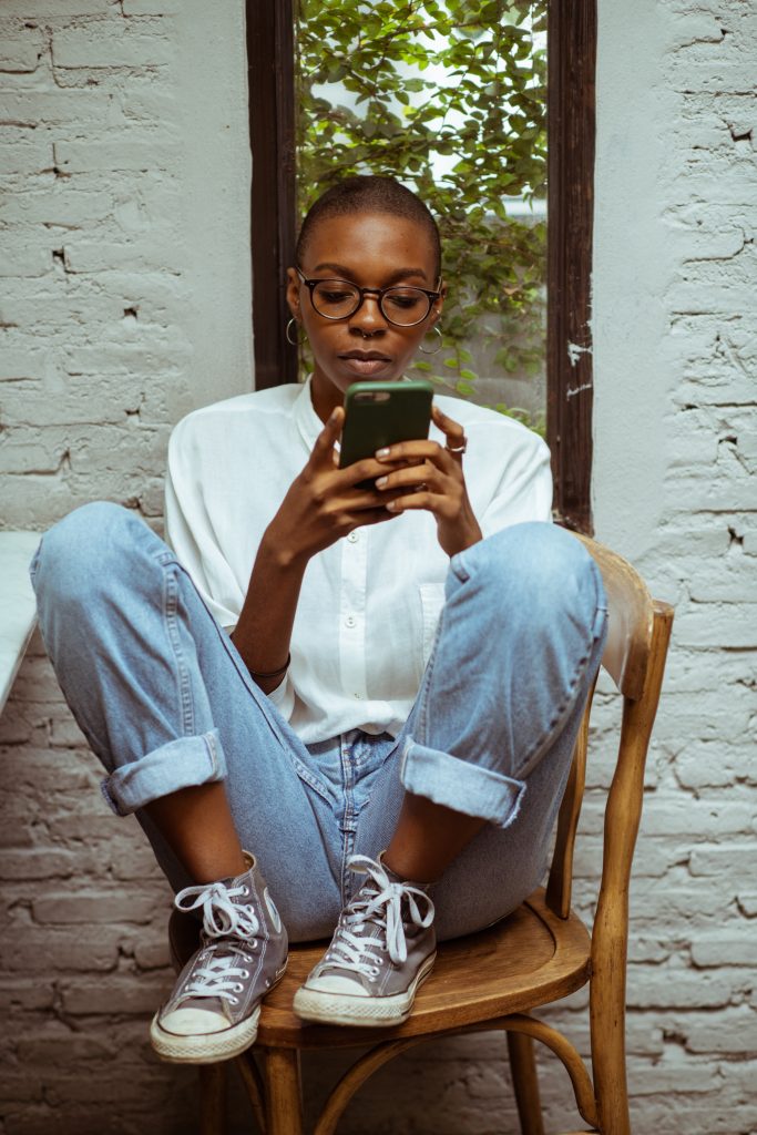 A Black women with a couple days of growth of a shaved head, wearing glasses, medium sized hoop earrings, a white collared button-down shirt, rolled up jeans and gray high top sneakers. She is looking at her phone while sitting on a cafe stool chair with her feet up on the chair and knees bent close to her chest. Behind her is a window and a brick wall, painted white.]