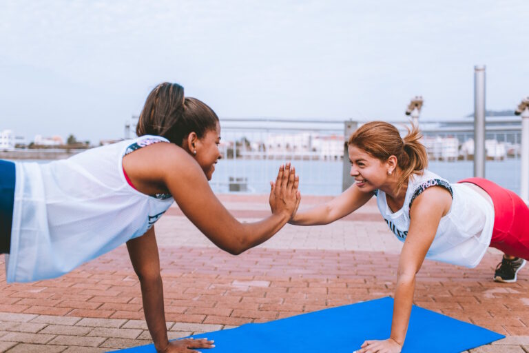 Two women face one another as they hold themselves up in plank positions and touch their palms together.