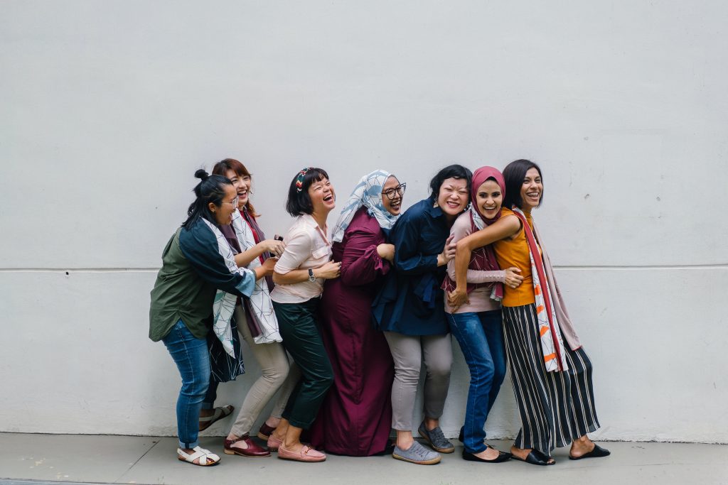 Seven young Asian women stand, leaning back into one another in a line. They are all smiling and laughing and wearing colorful clothes. Two of them are wearing hijabs.]