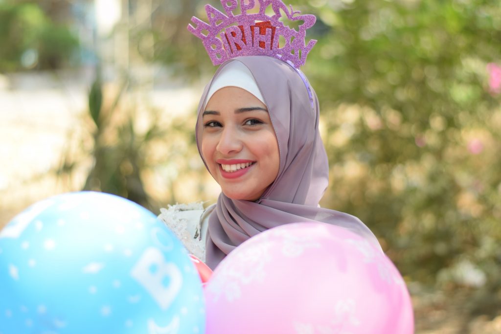 A teenage girl with olive skin, wearing a gray hijab with a happy birthday headpiece and balloons in front of her.]