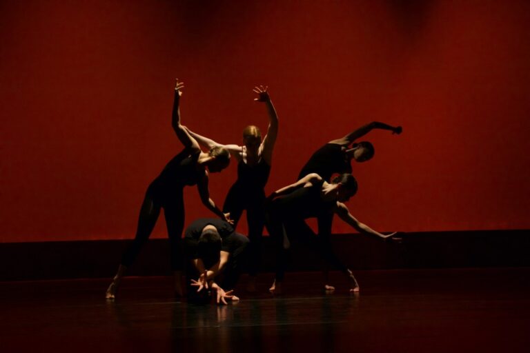 Five contemporary dancers on a dark stage with low lighting and a red background