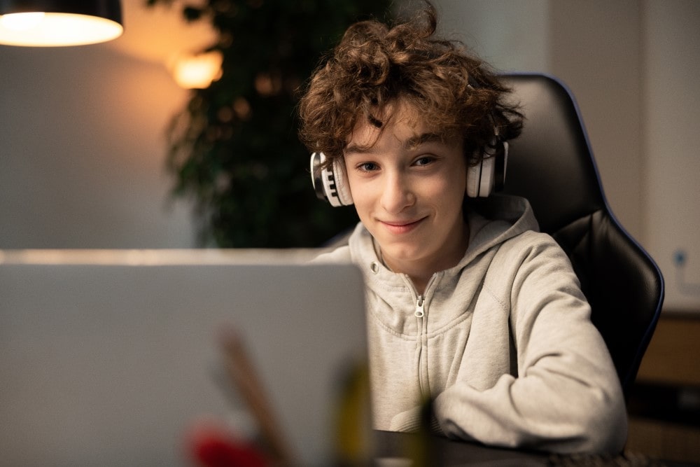 A smiling youth wearing headphones sitting in a gaming chair at a desk with a laptop computer