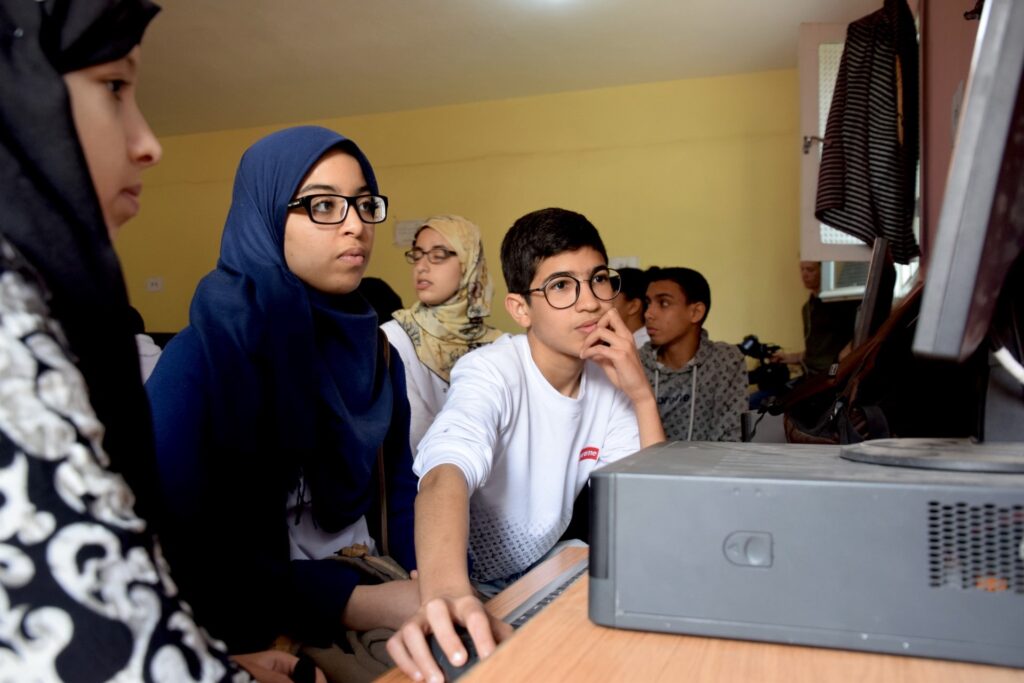 Two students, one wearing a headscarf, work on a desktop computer in a classroom
