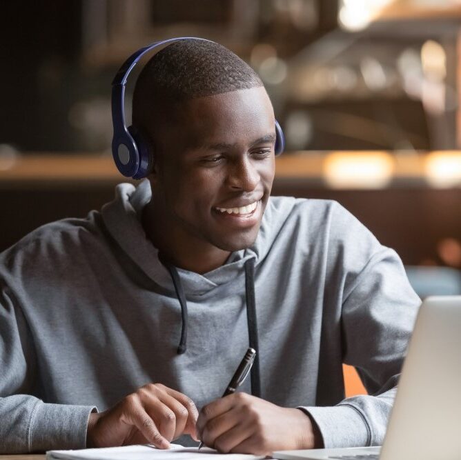 A smiling, dark-skinned youth in a sweatshirt and headphones takes notes with a pen and paper will using a laptop computer