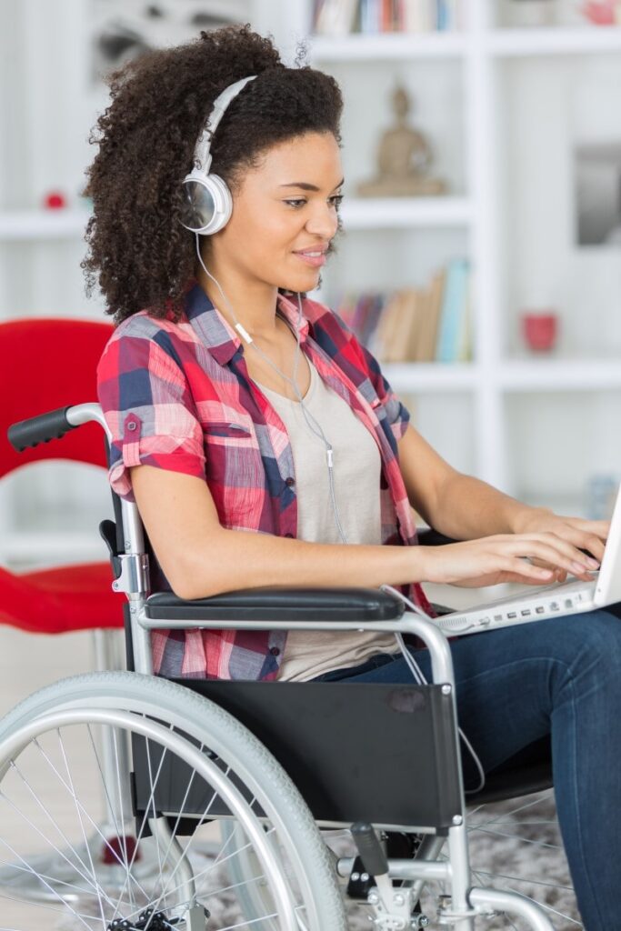 A woman with medium dark skin and curly hair in a wheelchair is working on a laptop computer while wearing a headset
