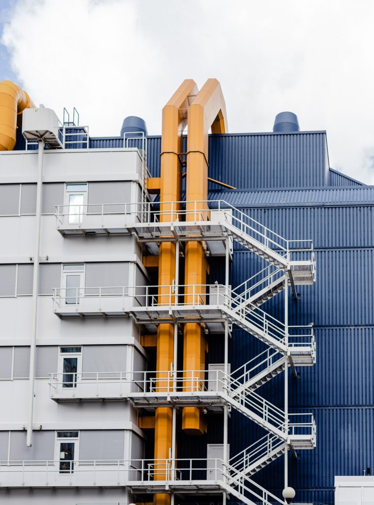 This image shows an industrial building with more than 4 floors colored grey and blue. It has 3 orange pipes extended to the last floor in addition to the outdoor stairs connecting each floor.