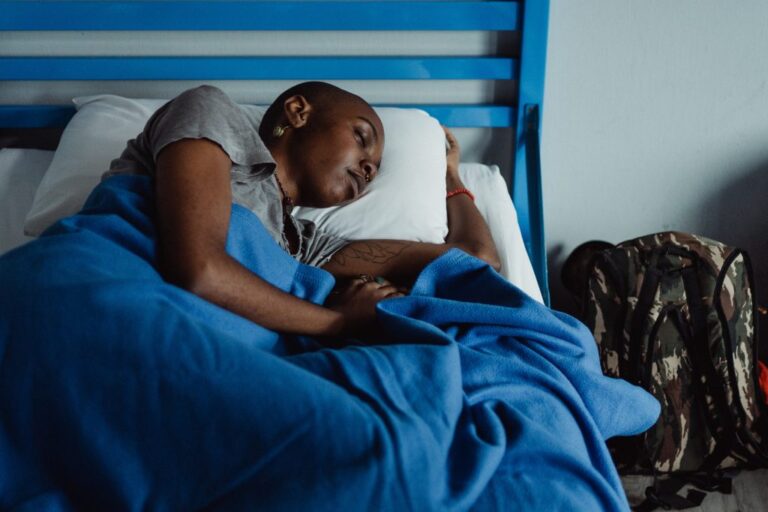 This image shows a dark-skinned young woman, head-shaved, wearing earrings, a septum piercing, an orange bracelet on her left biceps tattooed hand, a grey t-shirt, and sleeping on her metallic blue bed. She covers herself with a blue sheet next to a military backpack on her side.