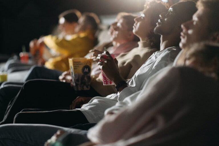 A variety of people -from different ages and races - are sitting on chairs, watching a movie, and having some popcorn in a dark room.