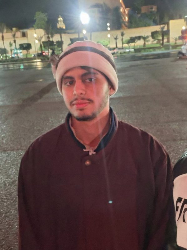 A young man with medium brown skin and a short black beard stands in a parking lot with a slight smile on his face. He is wearing a brown jacket and a striped hat.