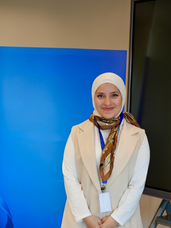 A smiling young woman stands with her hands clasped in front of a blue background and a TV screen. She is wearing a white hijab, a tan and white outfit, a colorful scarf, and a conference badge.