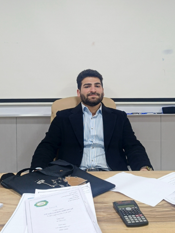 A young man with fair skin and a short black beard sits at a table in an office setting. He is wearing a blue and white striped shirt, a black blazer, and a slight smile on his face.