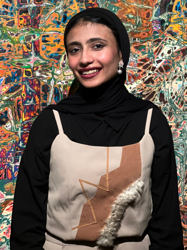A young woman with light brown skin smiles at the camera against a colorful background. She is wearing a black hijab, pearl earrings, and a tan patterned dress.