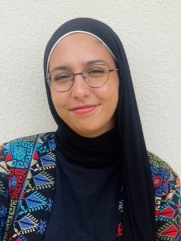 A young woman with light brown skin smiles at the camera against a white wall. She wears a colorful embroidered dress, a black hijab, and eyeglasses.
