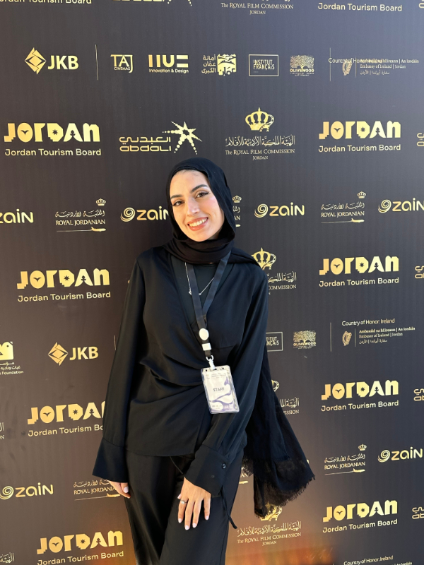 A smiling young woman in an all-black outfit and a black hijab stands in front of a black and gold event backdrop with various logos. She is wearing a ring, a necklace, and an event badge around her neck.