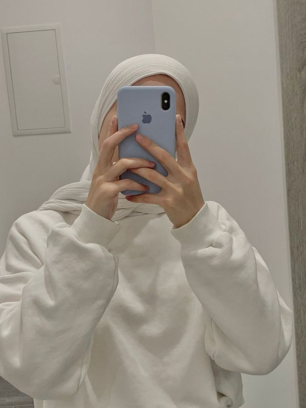 A young woman wearing a white hijab and a white hoodie takes a picture of her reflection in the mirror against a simple background, with a blue phone in front of her face.