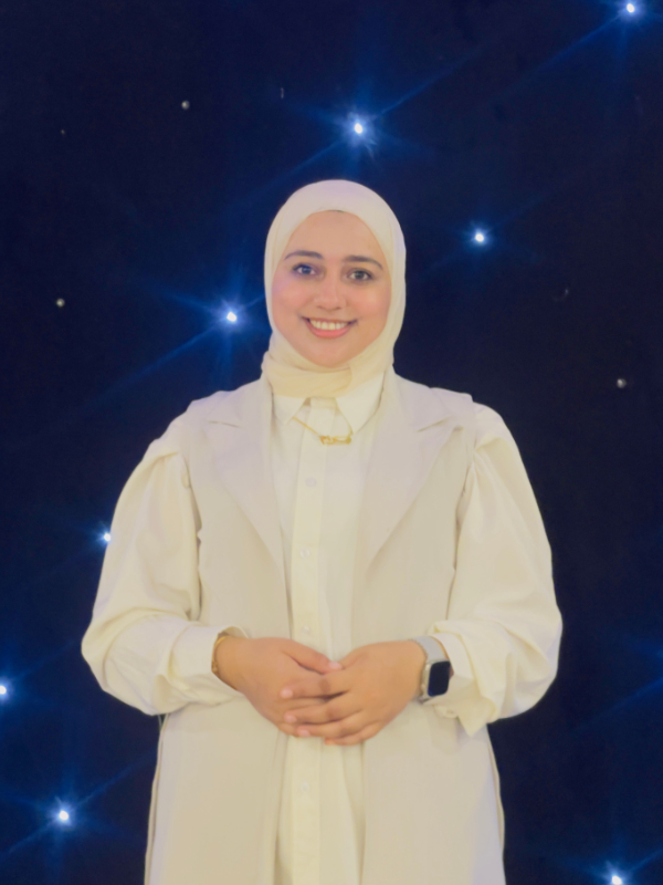 A young woman stands smiling with her hands clasped in front of her, against a dark-blue starry background. She wears an all-white outfit, a white hijab, a watch, and a gold necklace.