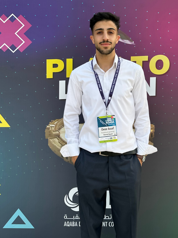 A young man with light brown skin, short black hair, and a short beard stands in front of a colorful event backdrop. He is wearing a white shirt, navy pants, a brown watch, and sporting dark black hair, a black beard, and a conference badge around his neck.
