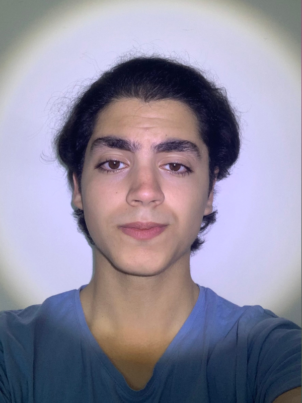A teen boy with fair skin, medium-length black hair, brown eyes, and thick eyebrows is wearing a blue shirt against a white background and has a neutral facial expression.