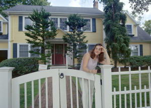 Young woman with pale skin and brown hair standing behind a white fence in front of a yellow house