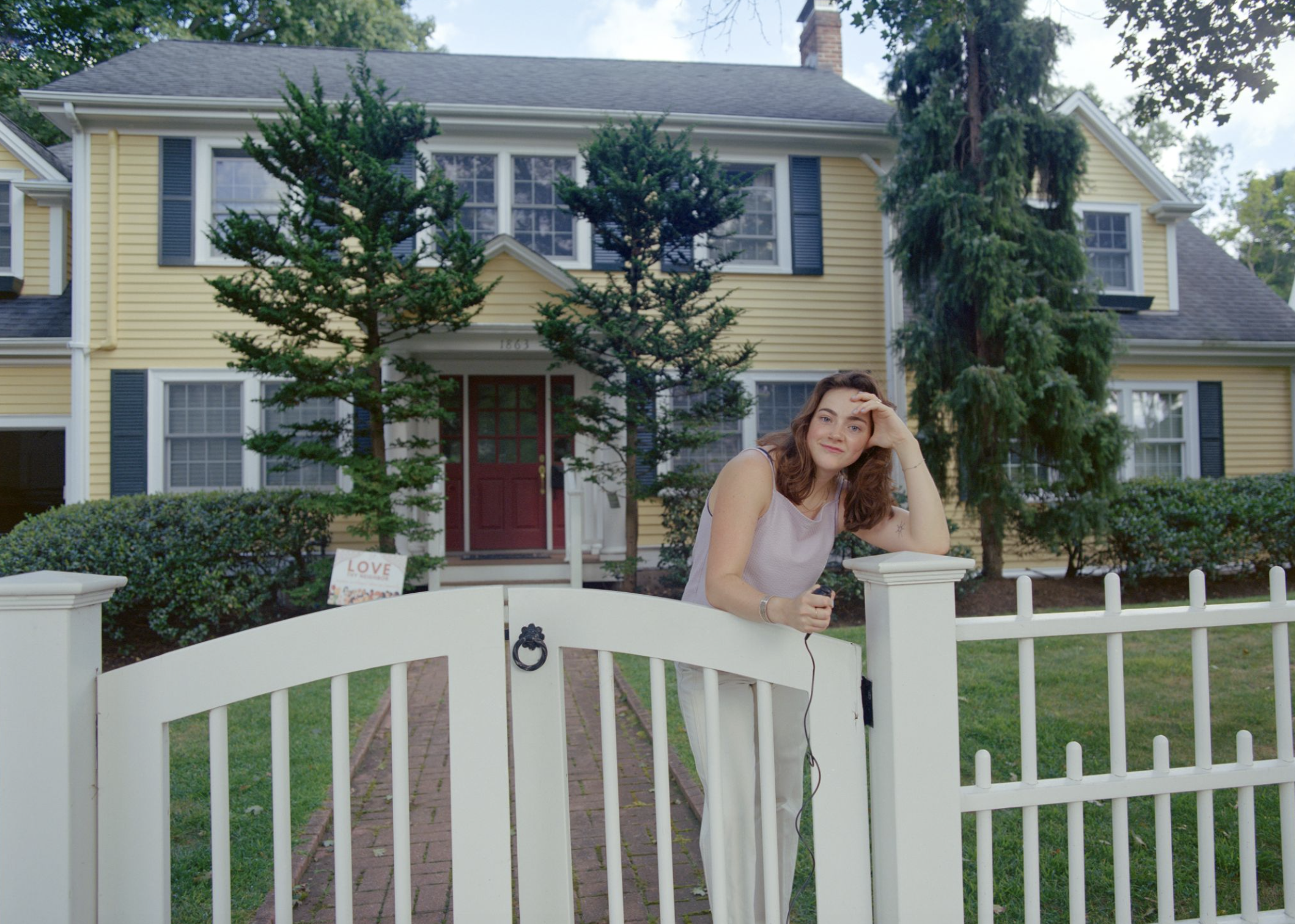 Young woman with pale skin and brown hair standing behind a white fence in front of a yellow house
