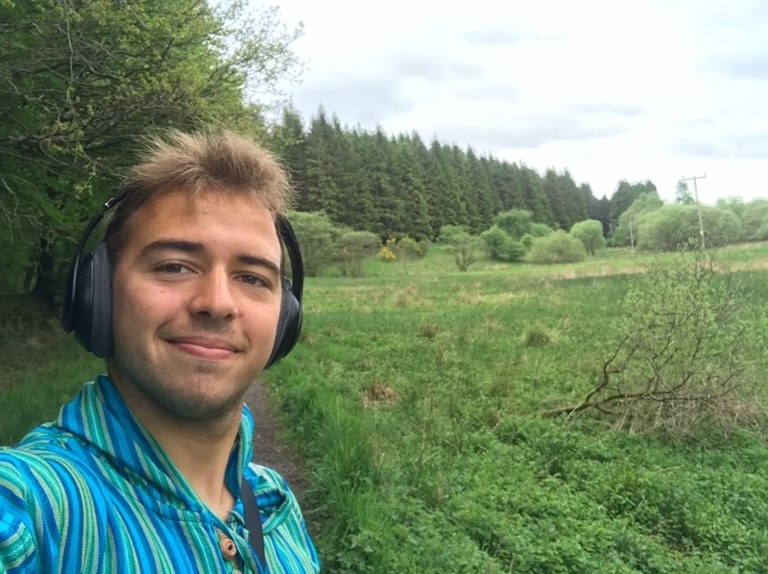 A young man smiling for a selfie while wearing large over-ear headphones and a teal and blue striped hooded shirt. He is standing on a path outdoors, with a wide green field and a dense forest of evergreen trees under a cloudy sky in the background.