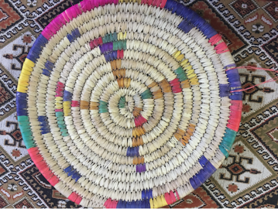 A top-down view of a circular, hand-woven straw basket featuring a spiral pattern. The basket is decorated with colorful accents in shades of pink, blue, yellow, and green, and is resting on a patterned rug.