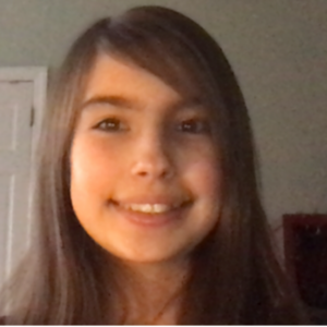 A young person with long brown hair and side-swept bangs smiling for a headshot. The background is a soft-lit indoor setting with a white door visible on the left.