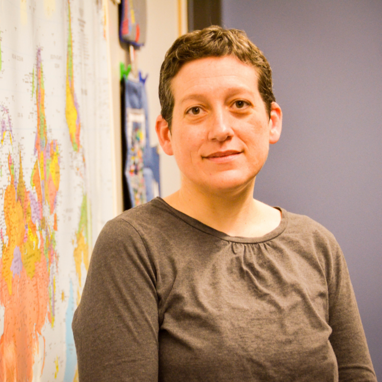 A woman with short, dark hair looking at the camera with a slight smile. She is wearing a grey long-sleeved shirt and standing in an indoor setting. Behind her is a colorful world map pinned to a wall, and a solid blue wall is visible behind her.