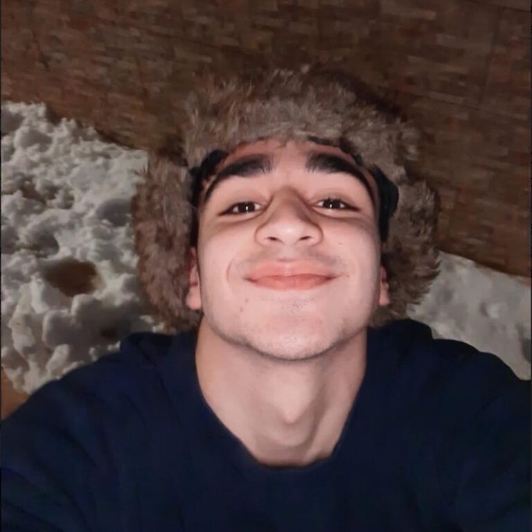 A young person with dark hair smiling for a close-up selfie while wearing a large, fuzzy brown faux-fur hat. He is wearing a dark blue shirt, and the background shows a textured brown wall and a patch of white snow.
