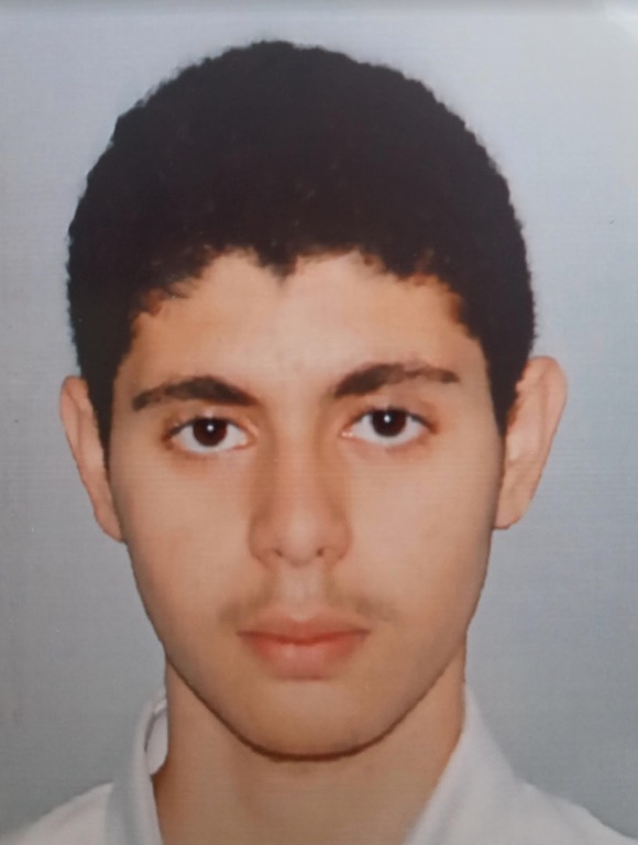 A young man with short, dark curly hair and dark eyes looking directly at the camera with a neutral expression. He is wearing a white collared shirt, and the photo is taken against a plain, light grey background.