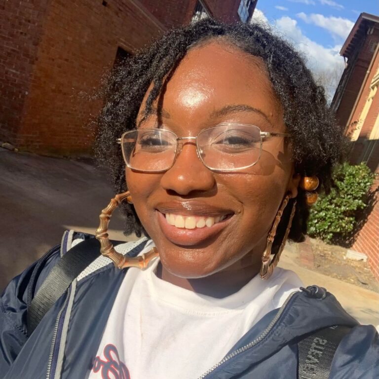 A smiling young woman with dark hair styled in short locs, wearing thin-rimmed glasses and large bamboo-style hoop earrings. She is wearing a white t-shirt under a dark jacket, posed outdoors in bright sunlight with a brick building and greenery in the background.