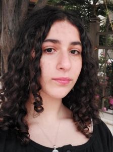 A close-up headshot of a young woman with long, dark curly hair looking at the camera with a soft expression. She is wearing a black top and a thin silver necklace with a pendant. The background is outdoors, featuring a tree trunk and a dark metal fence with pink flowers visible behind it.