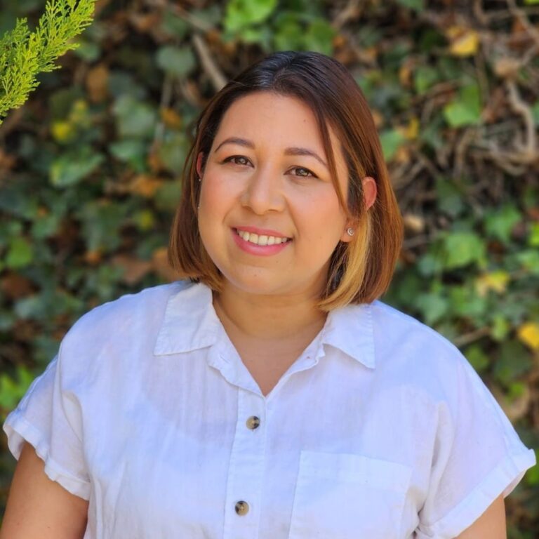 A smiling woman with shoulder-length brown hair featuring light blonde highlights. She is wearing a white short-sleeled button-down shirt and small stud earrings. The background is a soft-focus lush green hedge with hints of yellow leaves.