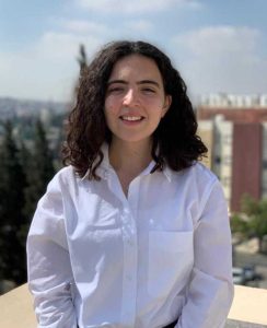 A young woman with shoulder-length curly dark hair smiling for a portrait. They are wearing a white button-down shirt and standing outdoors on a balcony or rooftop. The background shows a soft-focus view of a city with green trees and buildings under a bright, slightly cloudy sky.