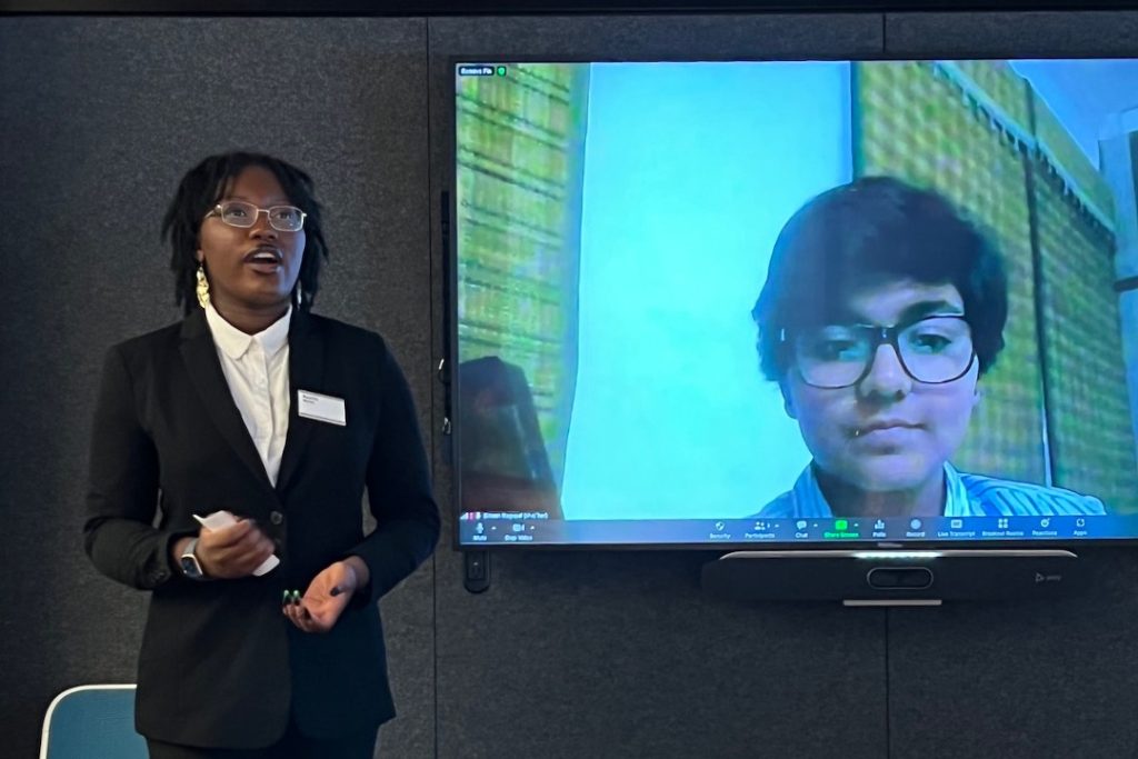 A person with dark hair and glasses wearing a black blazer and white shirt stands to the left, speaking while gesturing with their hands. To the right, a large television screen displays a video call with another person wearing glasses. The presentation is taking place in a room with dark grey textured walls.