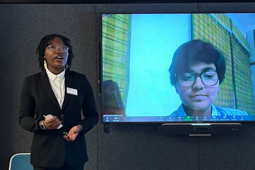A person with dark hair and glasses wearing a black blazer and white shirt stands to the left, speaking while gesturing with their hands. To the right, a large television screen displays a video call with another person wearing glasses. The presentation is taking place in a room with dark grey textured walls.